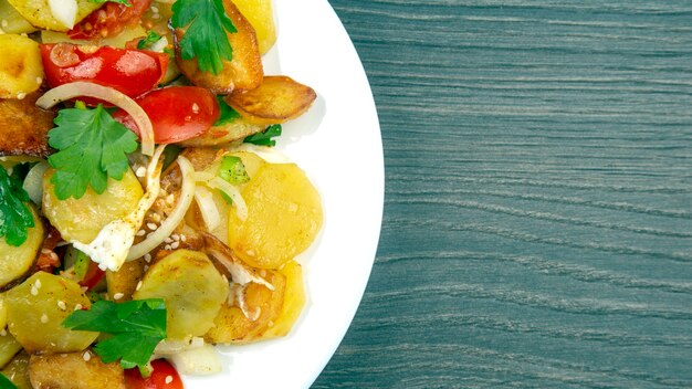 Cooked fried potatoes with herbs and vegetables in a white plate on a wooden table. copy space