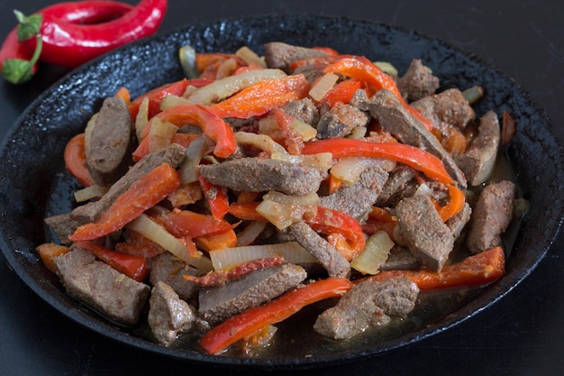 Cooked liver with vegetables on frying pan on black background