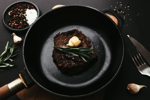 Cooked ribeye steak on cast-iron pan and board with pepper, rosemary, salt, garlic, knife, fork, butter on black