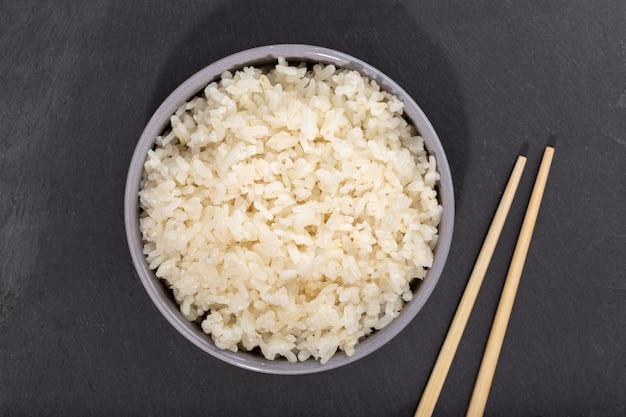 Cooked rice in bowl and chopsticks on black slate. Top view