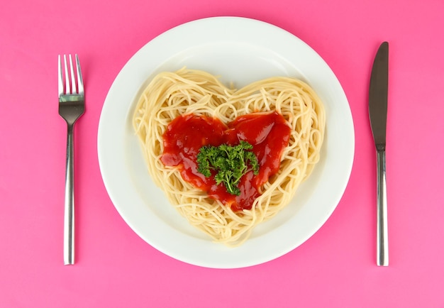 Приготовленные спагетти, тщательно уложенные в форме сердца и заправленные томатным соусом, на цветном фоне