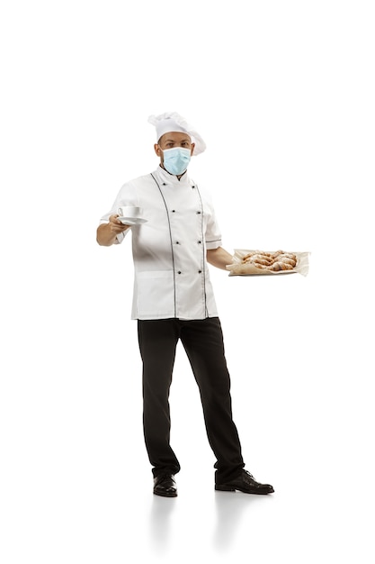 Cooker, chef, baker in face mask in uniform on white background with croissants. Young man, restaurant cooker's portrait. Business, foor, professional occupation, emotions concept. Copyspace for ad.