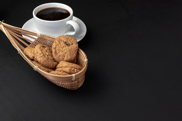 Cookies in bamboo basket and cup of coffee on black background