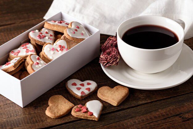 Cookies harten met suikerglazuur in witte doos en kopje rode thee en roos op houten achtergrond voor Valentijnsdag