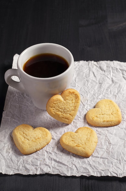 しわくちゃの紙にハートと一杯のコーヒーの形をしたクッキーは、黒い木製の背景にあります