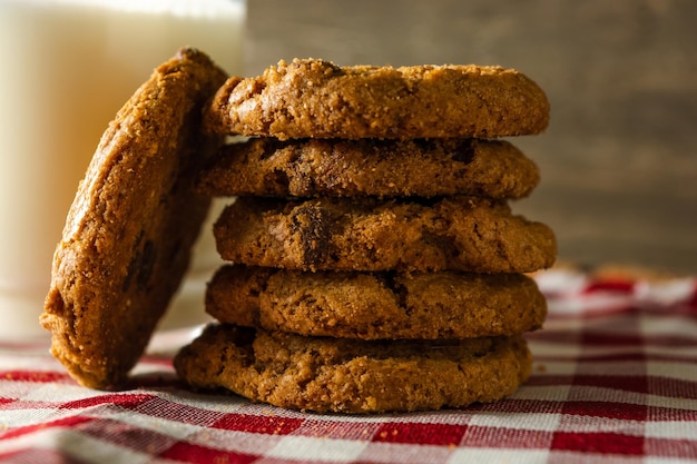 Cookies with chocolate drops stacked over each other and a\
glass of milk on red cloth table