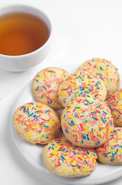 Cookies with colorful sprinkles in plate and cup of tea on white background close up