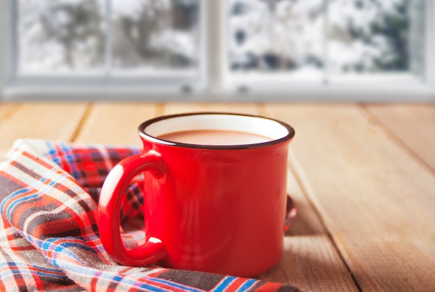 熱いお茶やコーヒーの木製のテーブルの赤いマグカップとクッキーの上の冷凍ウィンドウ。冬の暖かく居心地の良いコンセプト
