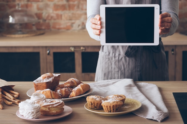 料理の広告。料理教室。黒い画面のタブレットを持つエプロンの男。焼きたてのケーキやペストリーの品揃え。