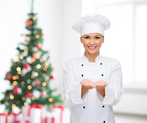 cooking, advertisement, holidays and people concept - smiling female chef, cook or baker holding something on palms of hand over living room and christmas tree background