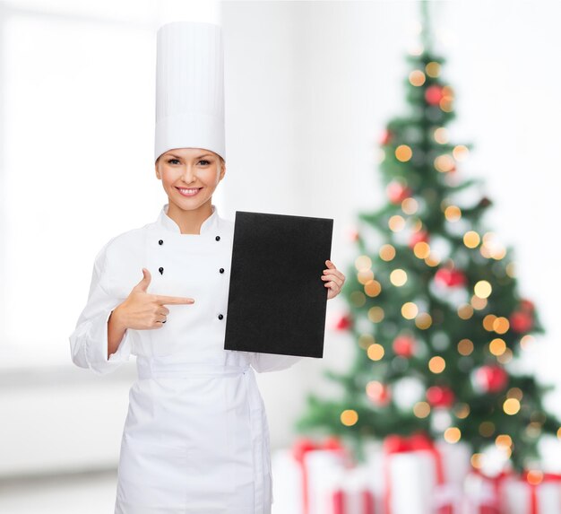 요리, 광고, 휴일 및 사람 개념 - 웃고 있는 여성 요리사, 요리사 또는 제빵사가 거실과 크리스마스 트리 배경 위에 있는 빈 검은색 메뉴 용지를 가리키는 손가락