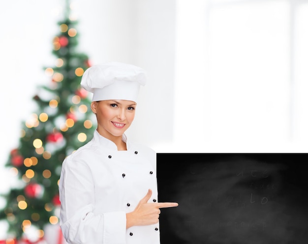 cooking, advertisement, holidays and people concept - smiling female chef, cook or baker pointing finger to blank blackboard over living room and christmas tree background