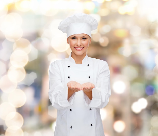 cooking, advertisement and people concept - smiling female chef, cook or baker holding something on palms of hands over holidays lights background