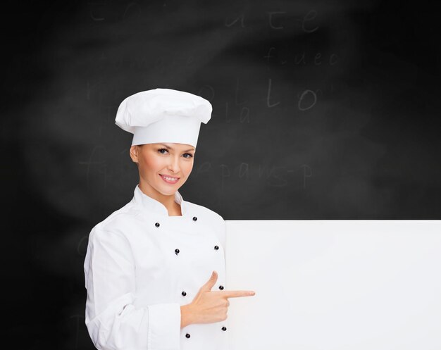 cooking, advertisement and people concept - smiling female chef, cook or baker pointing finger to white blank board over blackboard background