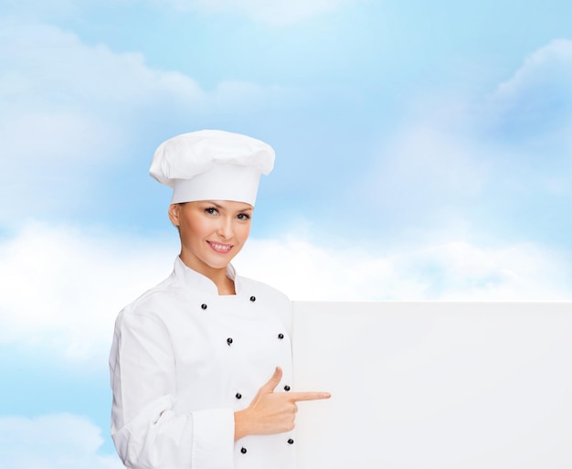 cooking, advertisement and people concept - smiling female chef, cook or baker pointing finger to white blank board over blue cloudy sky background