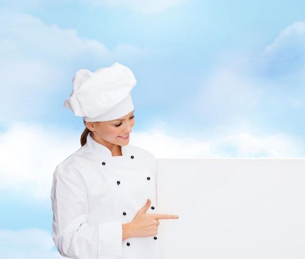 cooking, advertisement and people concept - smiling female chef, cook or baker pointing finger to white blank board over blue cloudy sky background