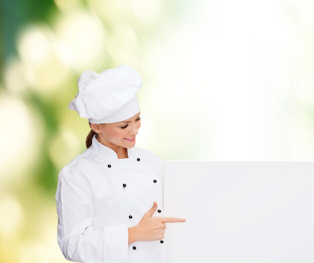 cooking, advertisement and people concept - smiling female chef, cook or baker pointing finger to white blank board over green background