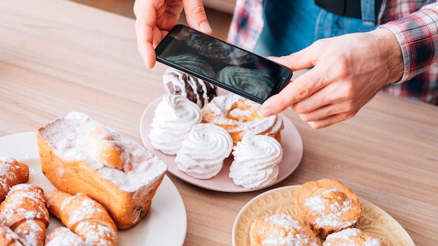 Кулинарный блог. Ассортимент тортов и выпечки. Мобильная фотография. Человек со смартфоном, снимающий сладкие хлебобулочные изделия.