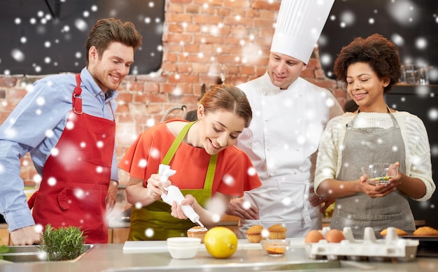 cooking class, culinary, bakery, food and people concept - happy group of friends and male chef cook baking in kitchen over snow effect