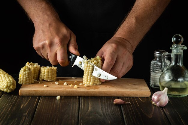 Cooking corn or maize grain by the hands of a chef with a knife on a wooden cutting board