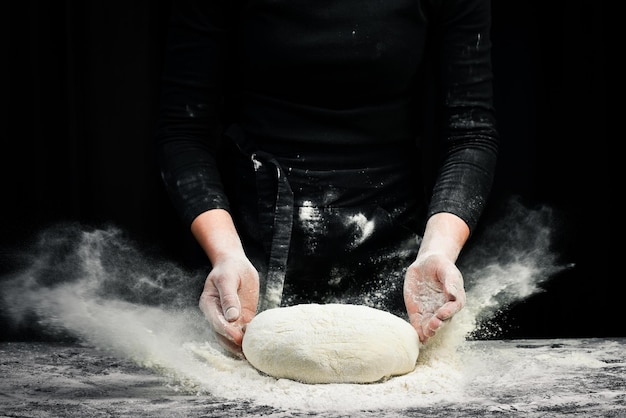 Cucinare l'impasto dalle mani dello chef per il pane di pasticceria fatto in casa con spruzzi di farina su uno sfondo scuro