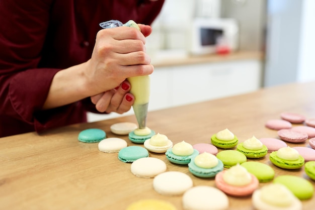 Photo cooking, food and baking concept - chef with confectionery bag squeezing cream filling to macarons shells at pastry shop kitchen