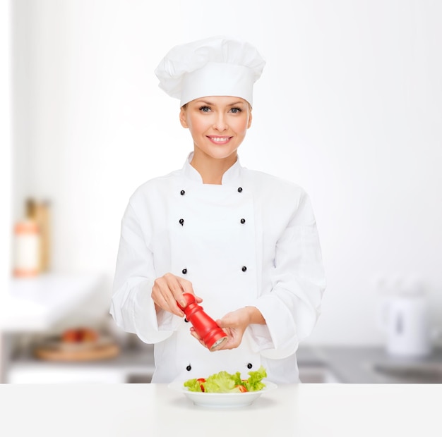 Concetto di cucina e cibo - chef femminile sorridente, cuoco o fornaio che prepara insalata con macinapepe o sale