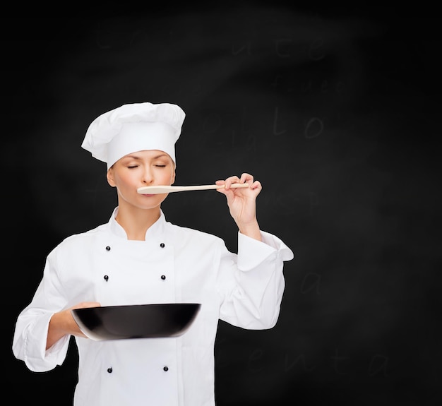 cooking and food concept - smiling female chef, cook or baker with pan and spoon tasting food