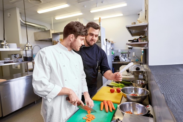 cooking food, profession and people concept - happy male chef cook with knife chopping carrot and looking to recipe at restaurant kitchen