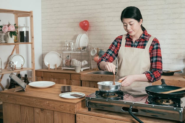 Cooking happy woman wear pinafore in wooden kitchen with pot\
stir melting chocolate to make sweet dessert for valentine day.\
young beautiful girlfriend handmade cocoa using spoon cooking on\
stove.