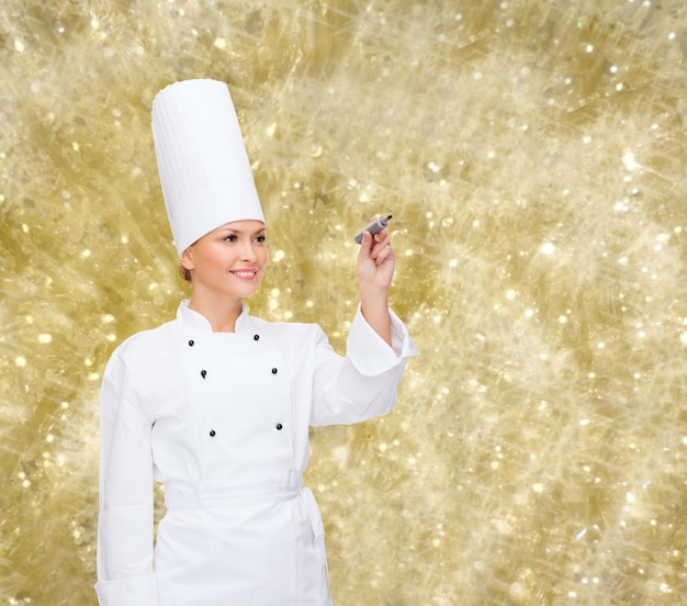 cooking, holidays, advertisement and people concept - smiling female chef, cook or baker with marker writing something on virtual screen over yellow lights background