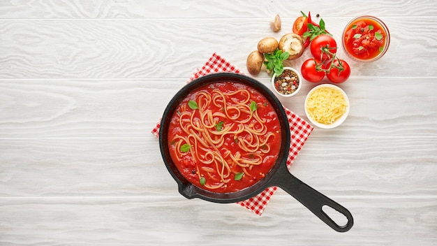 Cucinare la pasta fatta in casa con salsa di pomodoro in padella in ghisa servita con peperoncino, basilico fresco, pomodorini e spezie su un tavolo di legno di struttura bianca, concetto di cibo di ingredienti