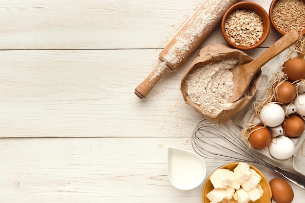 Cooking ingredients background. Border of flour, eggs, raisins, sugar, milk, butter and kitchen utensils on white rustic wood with copy space. Dough preparing and pastry concept, top view