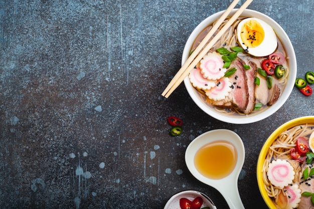 Cooking Japanese noodle soup ramen. Ramen with sliced pork, narutomaki, egg, kitchen spoon with broth on rustic stone background. Making traditional dish of Japan, top view, close-up. Space for text