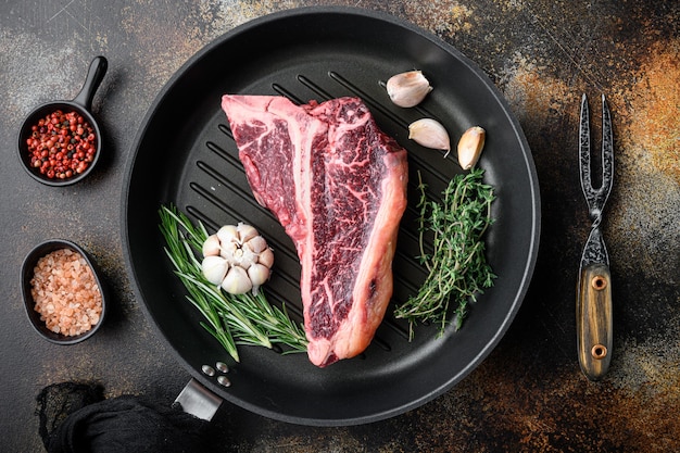 Sfondo di carne di cottura. bistecca con l'osso di manzo crudo invecchiato, con spezie ed erbe aromatiche per set grill, su padella in ghisa, su vecchio fondo rustico scuro, vista dall'alto distesa piatta