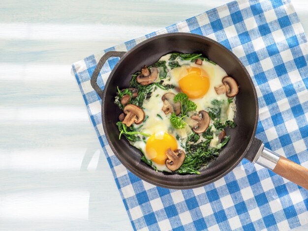 Cooking pan with spinach mushrooms cheese and fried eggs Healthy homemade dish for low carb diet on a light blue wooden table with linen towel Close up flat lay Top view Idea for breakfast