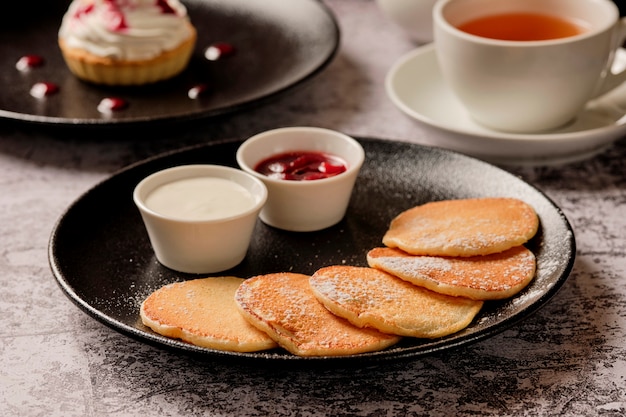 朝食のパンケーキを調理しています。サワークリームとベリージャムと背景に紅茶カップの小さな丸いフリッターをクローズアップ。