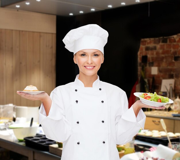 cooking, people and food concept - smiling female chef, cook or baker with salad and cake on plates over restaurant kitchen background
