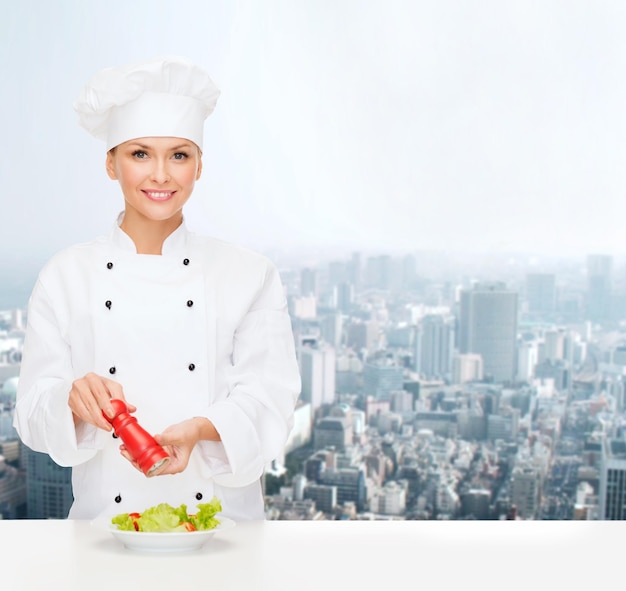 cooking, people and food concept - smiling female chef spicing vegetable salad over city background