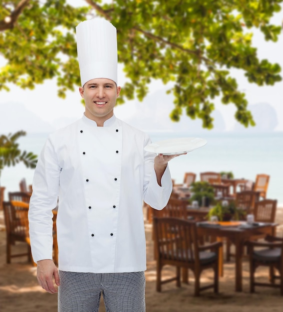 Foto concetto di cucina, professione, pubblicità e persone - cuoco maschio felice che mostra qualcosa sul piatto vuoto sopra la lounge del ristorante sulla spiaggia