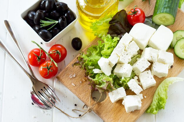 Приготовление салата из ручья со свежими овощами, сыром фета и маслинами на белом деревянном столе