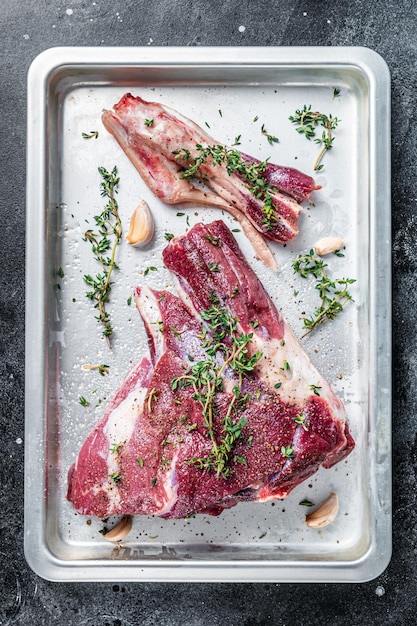 Cooking of Raw lamb mutton leg with thyme, pepper and olive oil in baking tray. Black background. Top view.