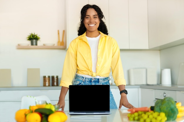 Cooking recipes online happy black lady showing laptop with blank screen in modern kitchen mockup copy space