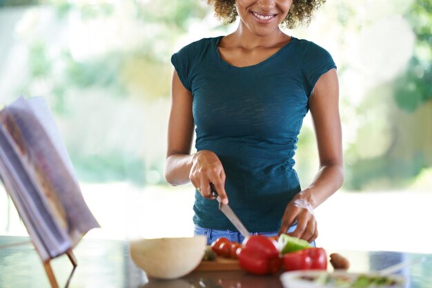 Foto cucinare sorriso e mani tagliare verdure in cucina per una dieta sana nutrizione o pranzo tagliare cibo a tavola e persona felice preparare insalata per cena o pasto biologico con libro di ricette a casa