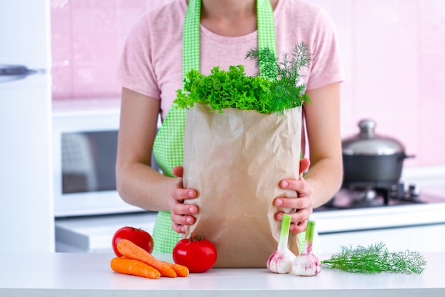 キッチンで新鮮な有機野菜のクラフトペーパーバッグを保持しているエプロンで女性を調理します。健康食品とバランスの取れた食事