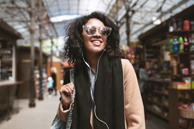 Cool African American girl standing on street in earphones. Portrait of beautiful girl with dark curly hair in sunglasses walking around city