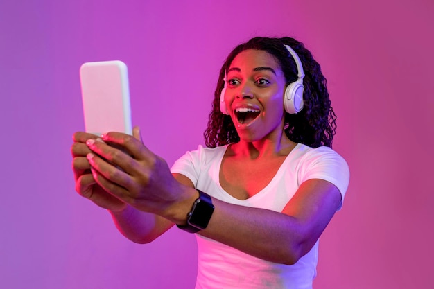 クールなアプリはスマートフォンの画面を見てワイヤレス ヘッドフォンを身に着けている黒人女性を驚かせた
