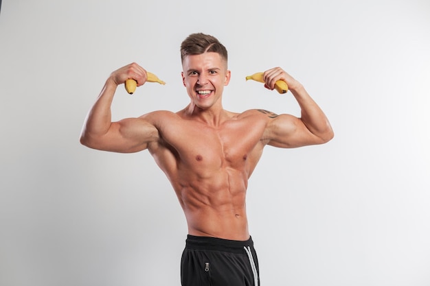 Cool athletic handsome funny sports man with emotions with a muscular body holding bananas and showing biceps on a white background in the studio Raw food and vegan