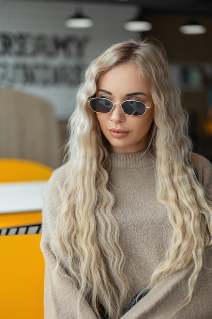 베이지색 스웨터에 빈티지 선글라스를 쓴 금발 곱슬머리 스타일의 멋진 젊은 패션 여성 모델이 카페에 앉아 있습니다.