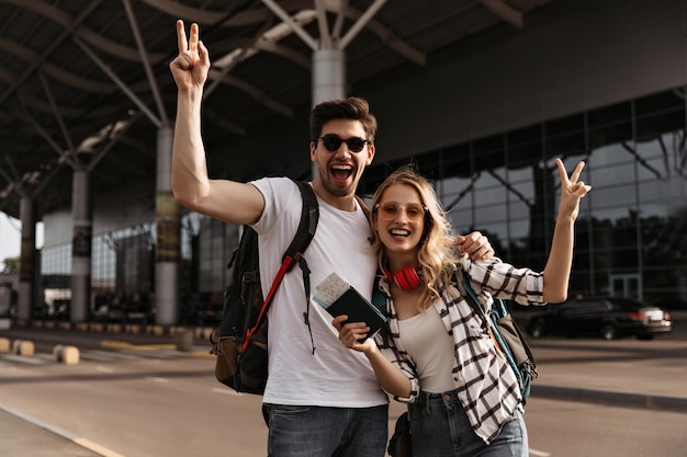 Cool blonde woman and brunette man in white tee shows peace signs on airport background Charming girl in plaid shirt and guy in white tee poses with backpacks passport and plane tickets